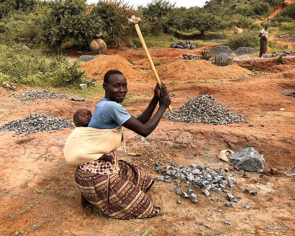 A Ugandan woman swings a sledge hammer at a pile of rocks while a baby sleeps peacefully on her back.